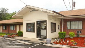 A small, single-story motel with a beige exterior and a black door. There are flower beds with red mulch and various plants in front of the building.