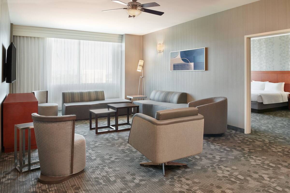A hotel room with a sofa, armchairs, a coffee table, a TV, a ceiling fan, and a bed.