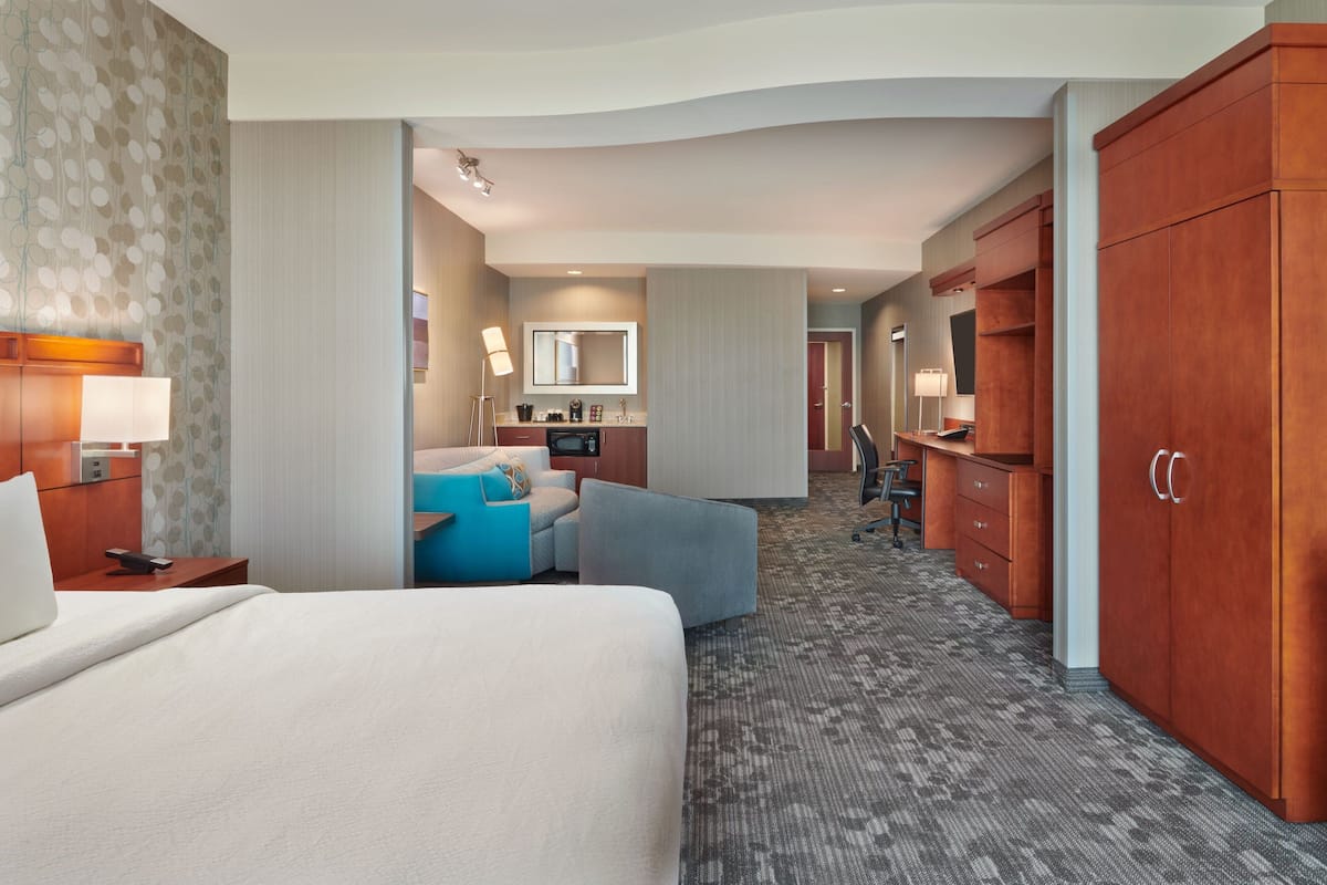 A hotel room with a large bed, a desk with a lamp, a sofa, a bathroom with a mirror, and a wardrobe.