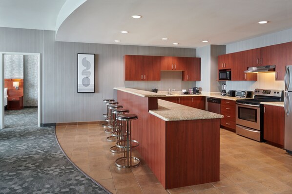A modern kitchen with wooden cabinets, a central island with bar stools, stainless steel appliances, and a tiled floor.
