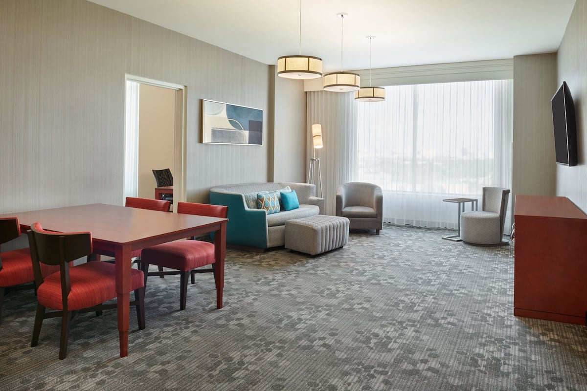 A hotel room with a dining area, a sofa set, a chair, a TV mounted on the wall, and a large window with curtains.