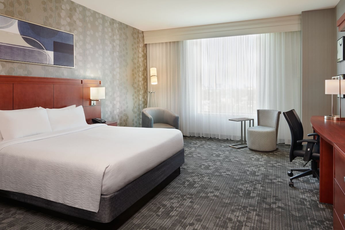 A hotel room with a large bed, a desk, two chairs, a small table, and a large window with curtains.