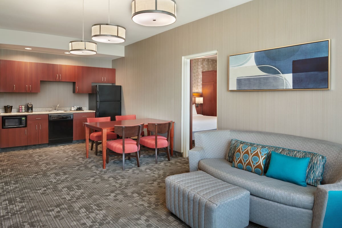 A modern hotel room with a kitchenette, a dining area with red chairs, a grey sofa with patterned cushions, and a large abstract painting on the wall.