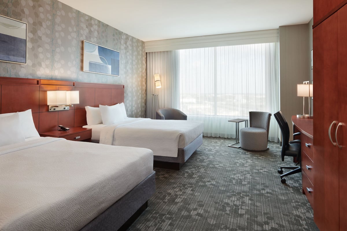 A hotel room with two beds, a desk, a chair, a wardrobe, and a large window with curtains.