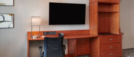 A hotel room with a wooden desk, a flat-screen TV mounted on the wall, a desk lamp, a chair, and a small drawer unit.