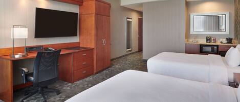 A hotel room with a desk, two beds, a TV, a mirror, and a kitchenette.
