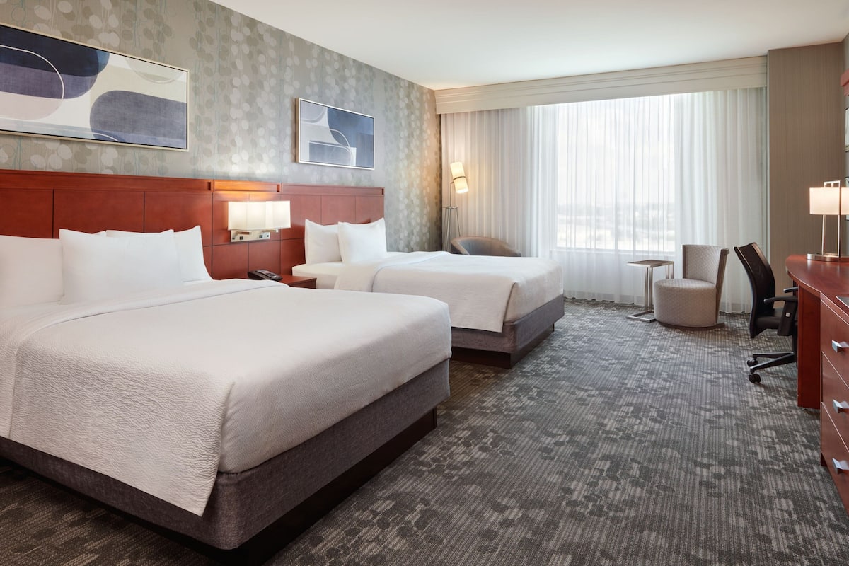 A hotel room with two beds, a desk, a chair, and a large window with curtains.