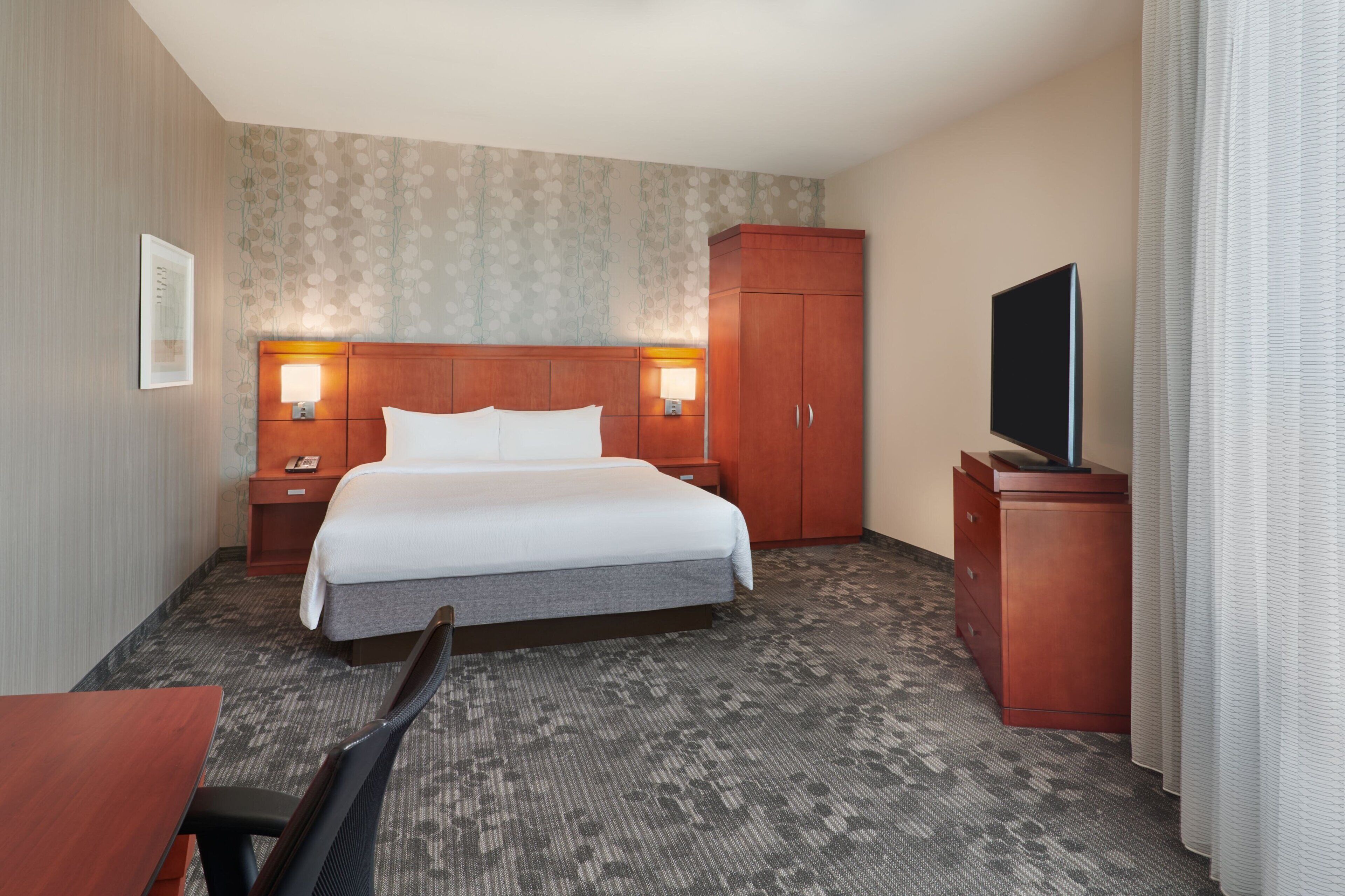 A hotel room with a large bed, a wooden wardrobe, a desk with a chair, a television, and a patterned carpet.