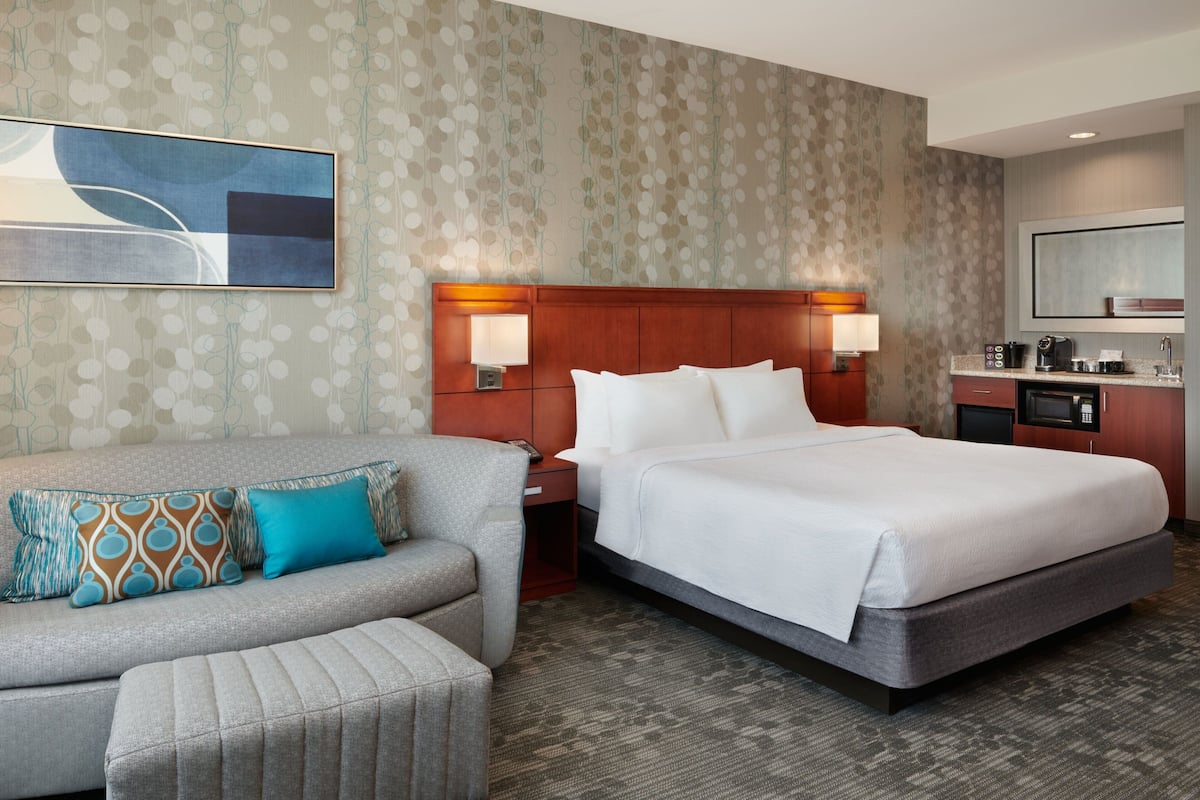A hotel room with a large bed, a sofa, a nightstand, a desk, and a kitchenette.