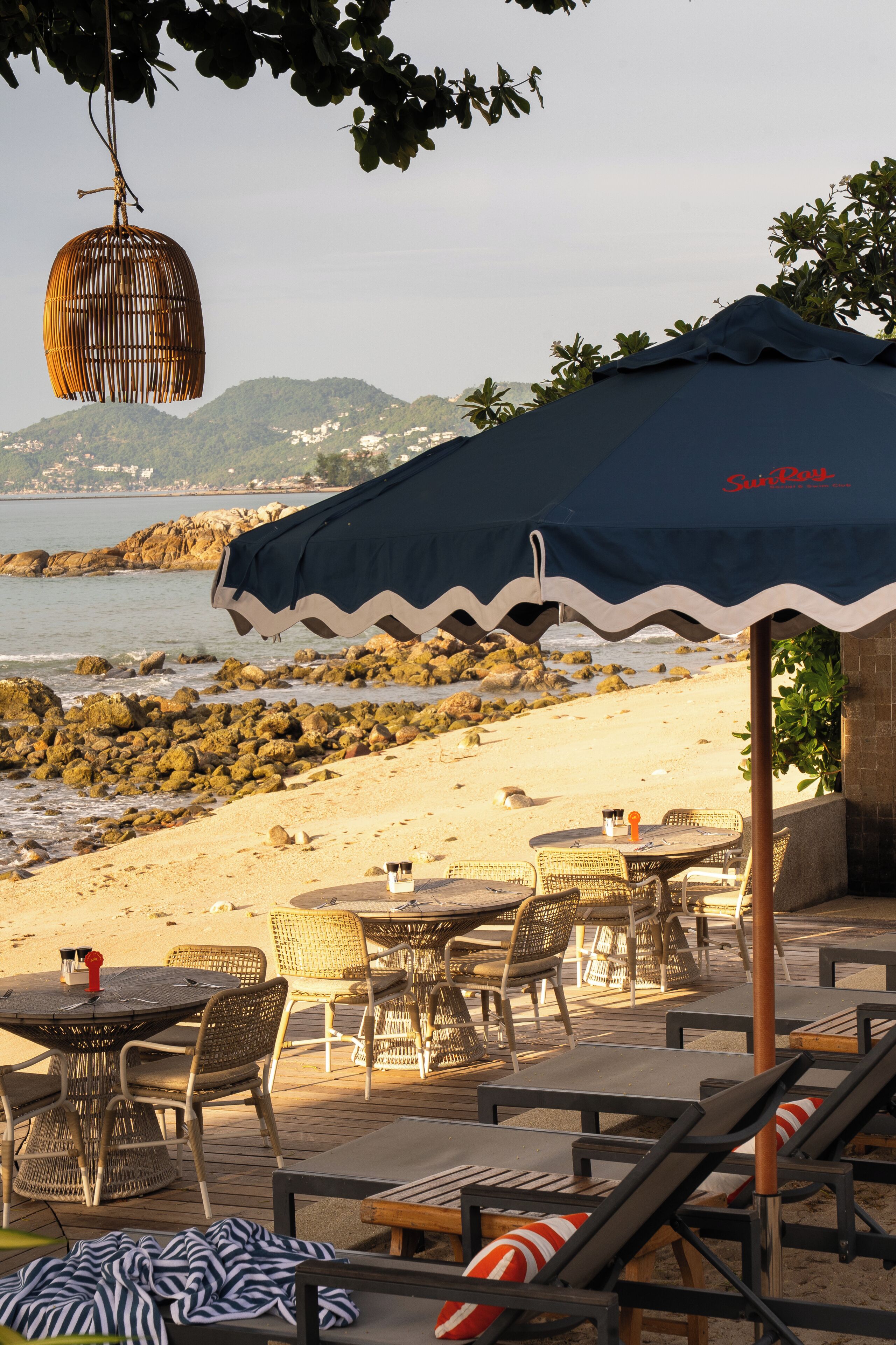 Un área exterior con mesas y sillas, una lámpara colgante de mimbre y vistas a la playa y al océano.
