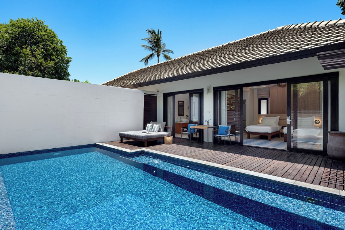 A modern pool area with a clear blue pool, a wooden deck, and a bedroom with a bed and seating area.