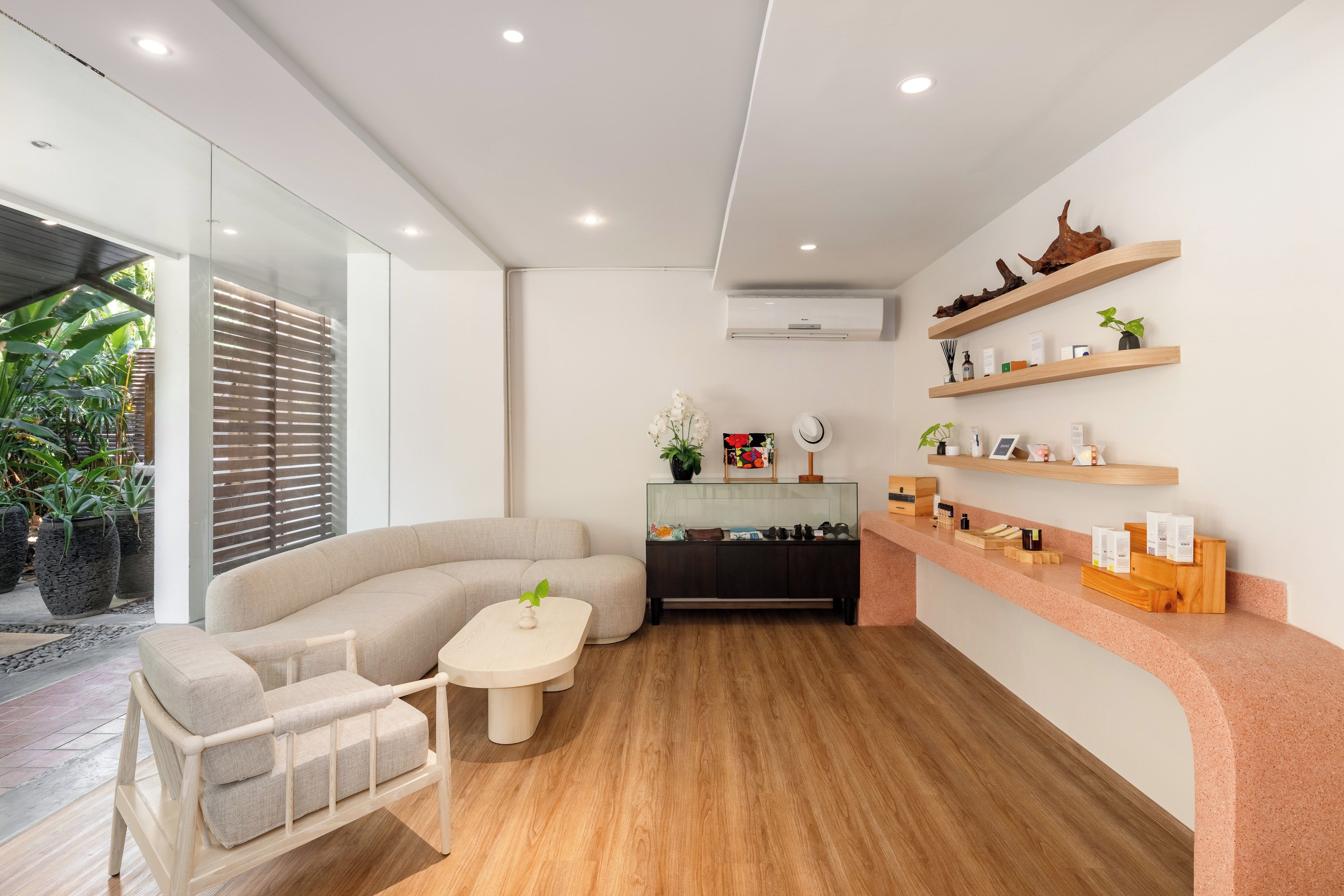 A modern living room with a curved sofa, a small table, and a wooden floor. There is a glass partition on one side and a wooden shelf with various items on the other.