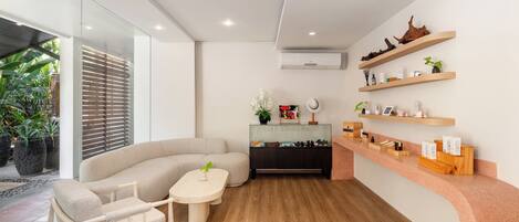 A modern living room with a curved sofa, a small table, and a wooden floor. There is a glass partition on one side and a wooden shelf with various items on the other.