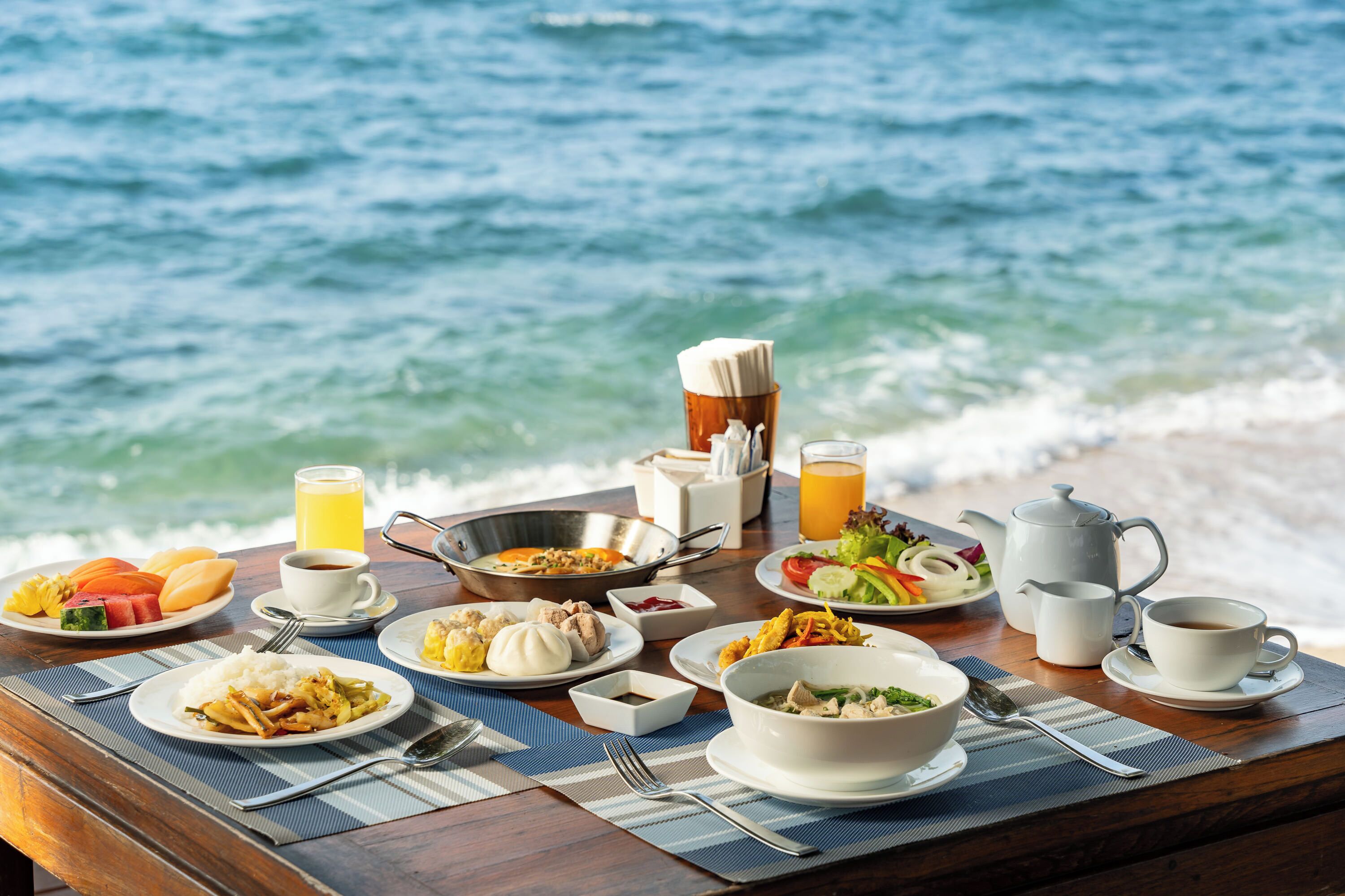 A table set with various dishes, drinks, and a teapot, overlooking the sea.