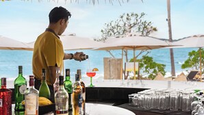 A bar area with a bartender preparing a drink, bottles of alcohol, and glasses on the counter, overlooking a beach. - SAii Koh Samui Villas - Adults Only (Koh Samui)