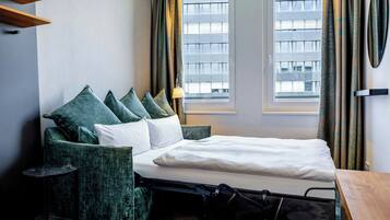 Quarto de hotel com cama, cadeira, luminária e janela com cortinas.