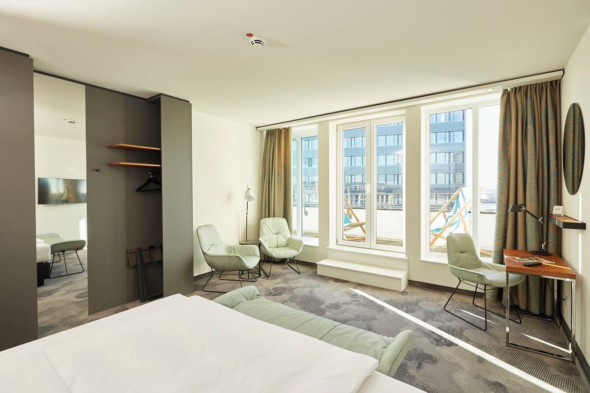 A modern hotel room with a large bed, two armchairs, a desk, and a view of the city through large windows.