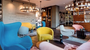 Un moderno espacio de descanso con diversas opciones de asiento, incluyendo un sillón amarillo, un sillón azul y una silla rosa. Hay varias estanterías llenas de libros y objetos decorativos.