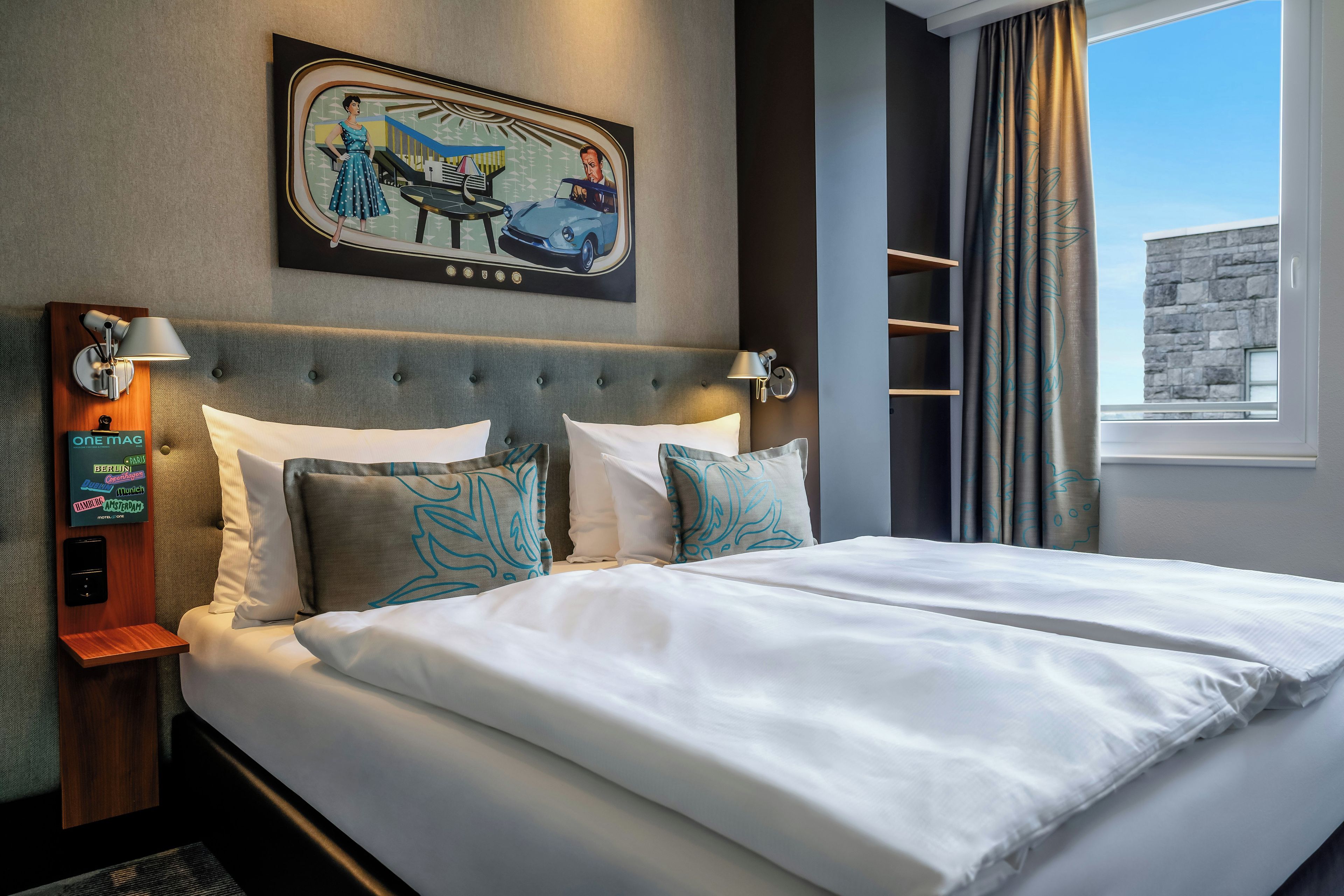 A hotel room with a bed, a wall-mounted artwork, a nightstand, a lamp, and a window with curtains.