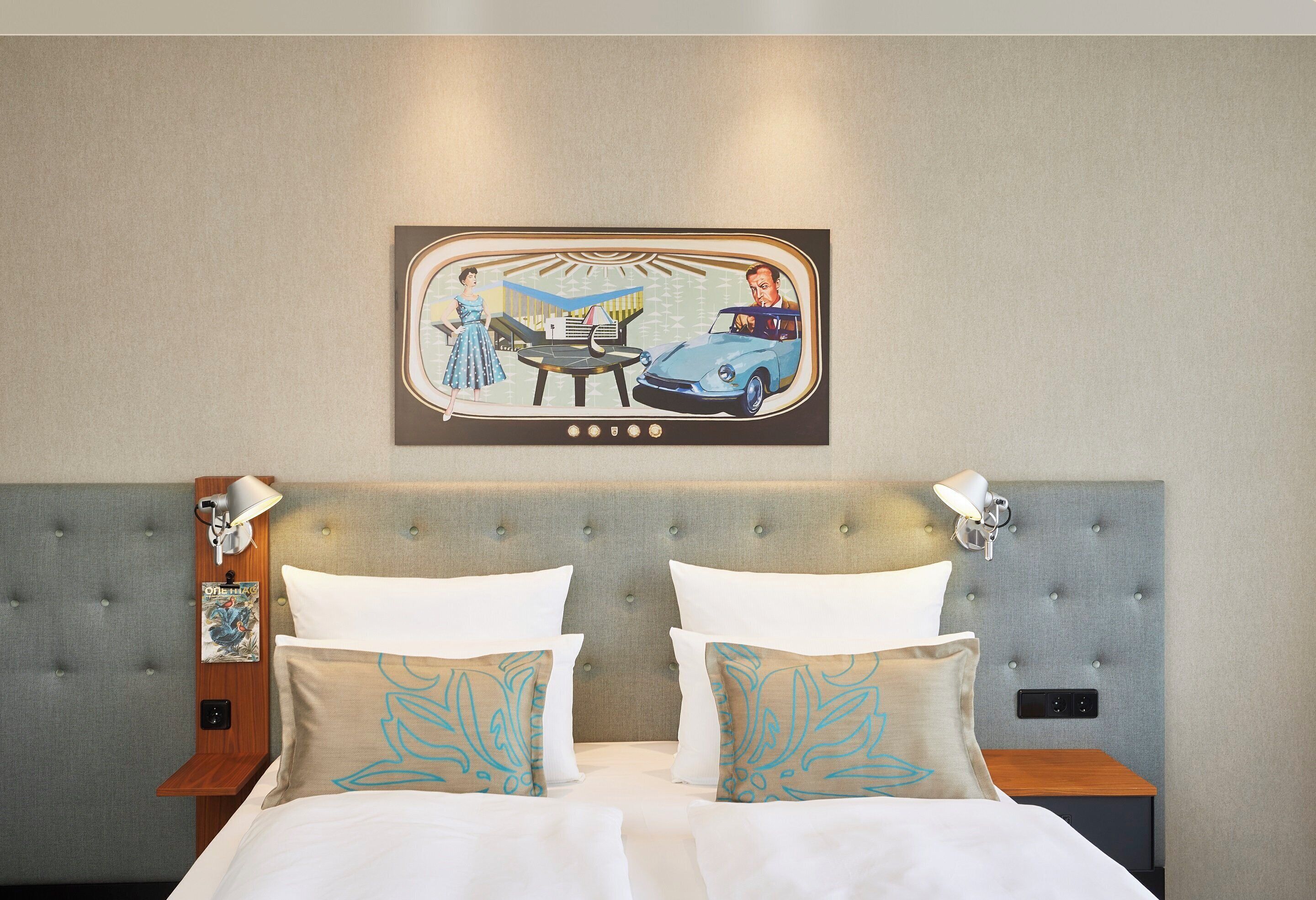 A hotel room with a tufted headboard, two bedside tables with lamps, a bed with white linens, and a wall-mounted picture of a vintage car scene.