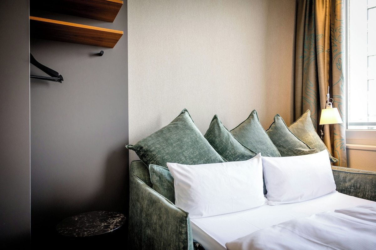 Una habitación de hotel con una cama, almohadas, una lámpara y un ventanal con cortinas.