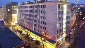 Un edificio moderno con una fachada de ventanas enrejada, techo plano y estacionamiento con coches.