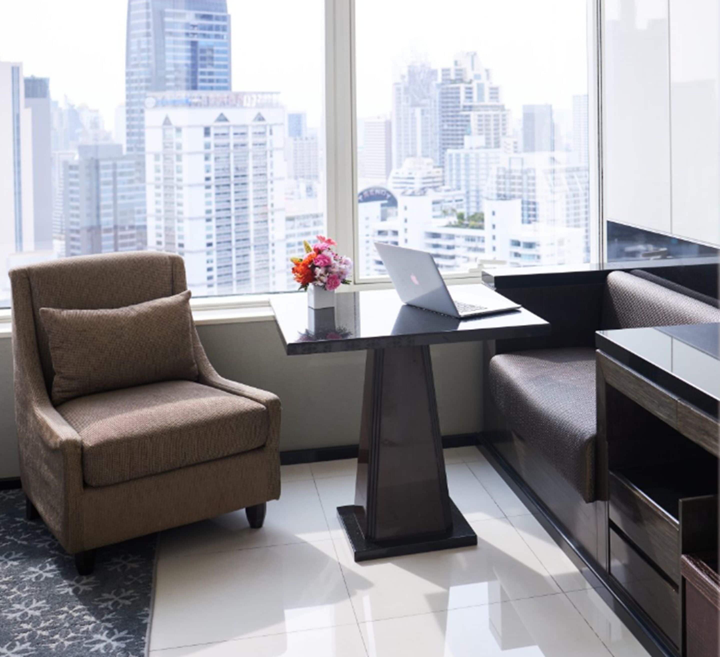A modern office space with a sofa, a desk with a laptop, and a view of a cityscape through large windows.