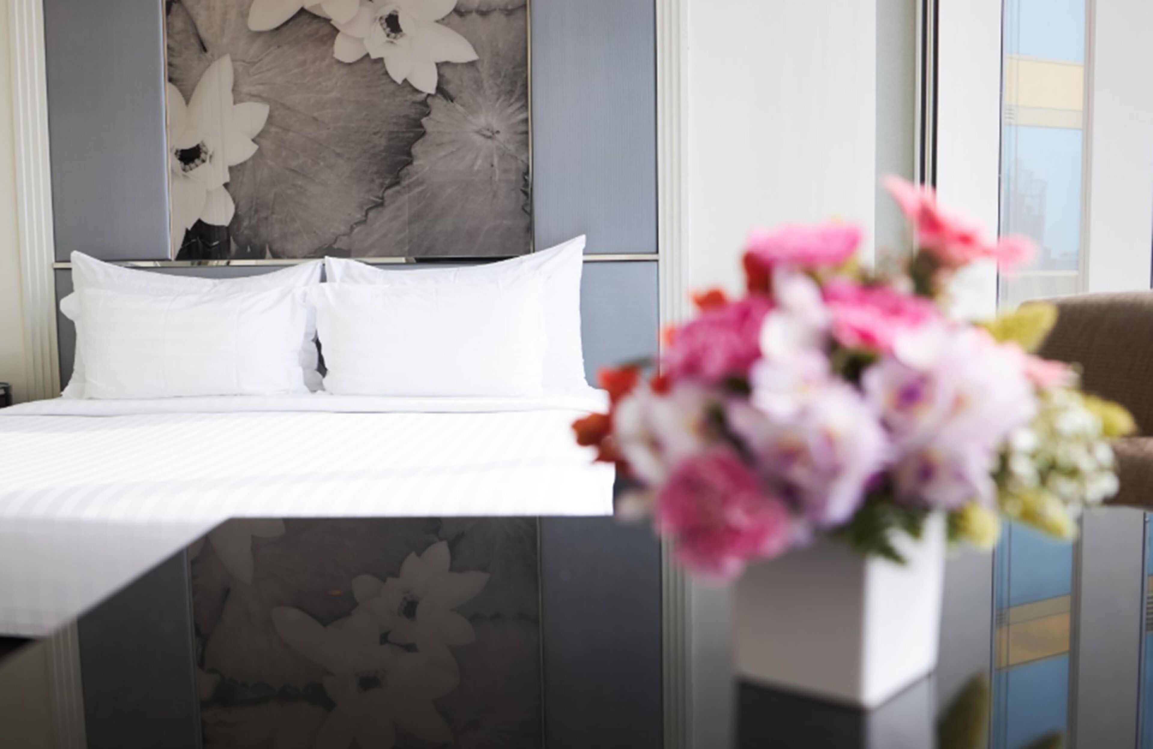 A hotel room with a bed, white pillows, a floral wall decoration, and a vase with flowers.