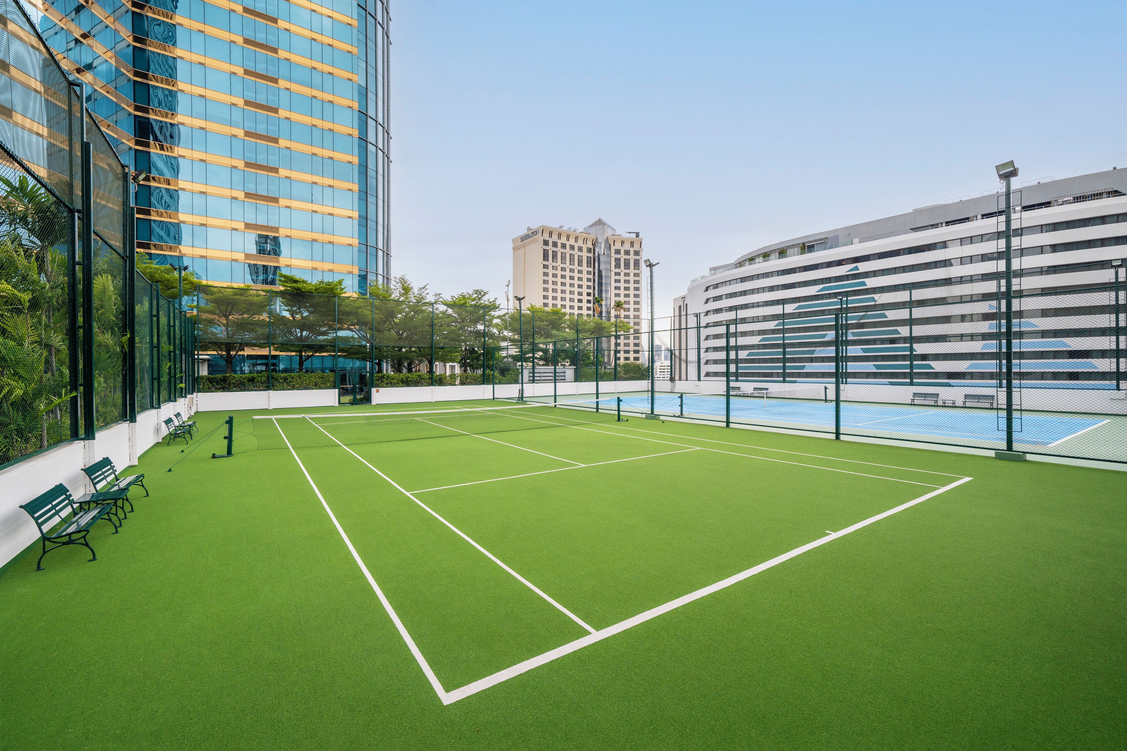 An indoor tennis court with green artificial turf, surrounded by modern buildings and a pool area.