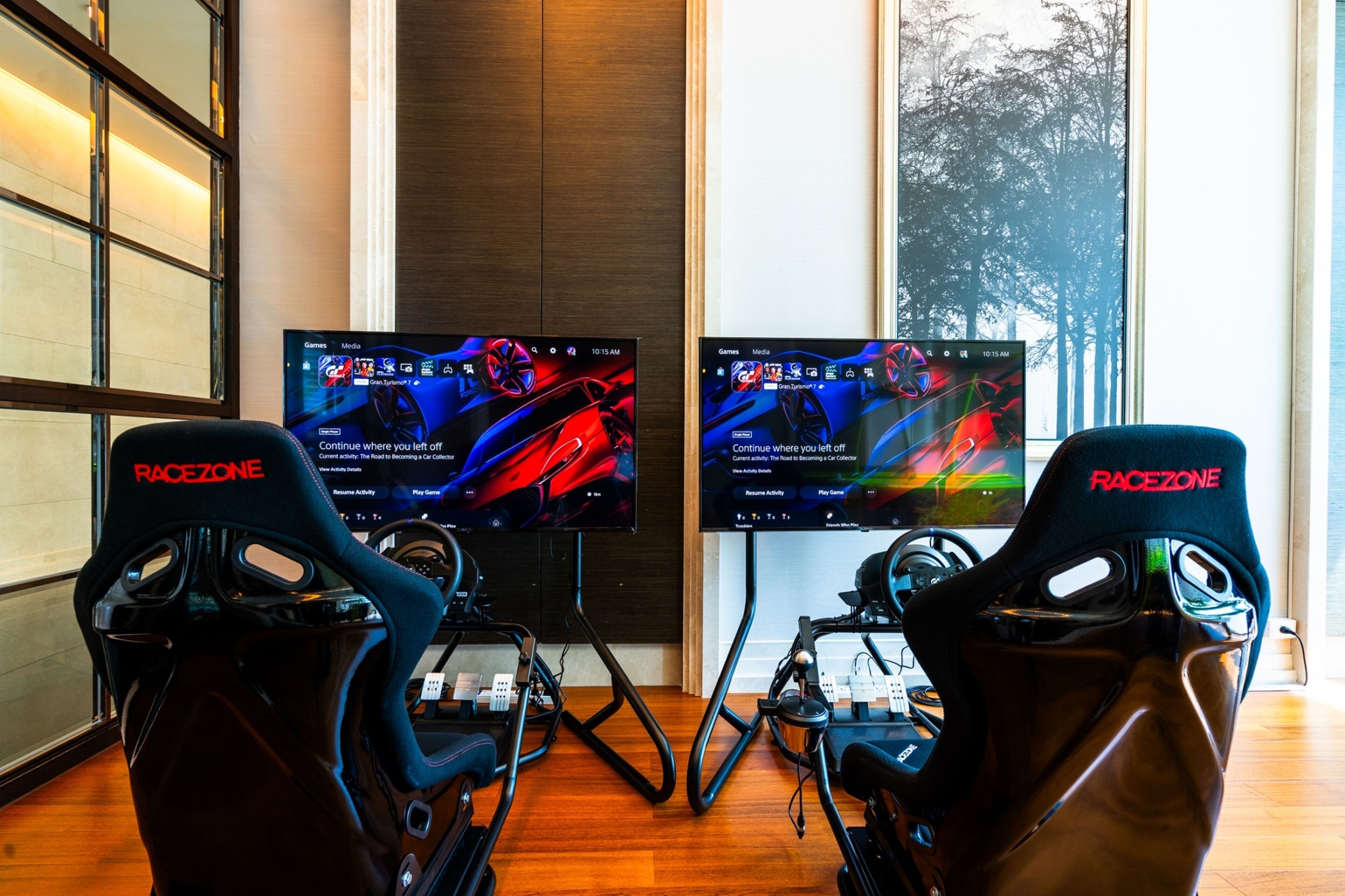 A gaming setup with two monitors displaying a racing game, two racing chairs, and a wooden floor.
