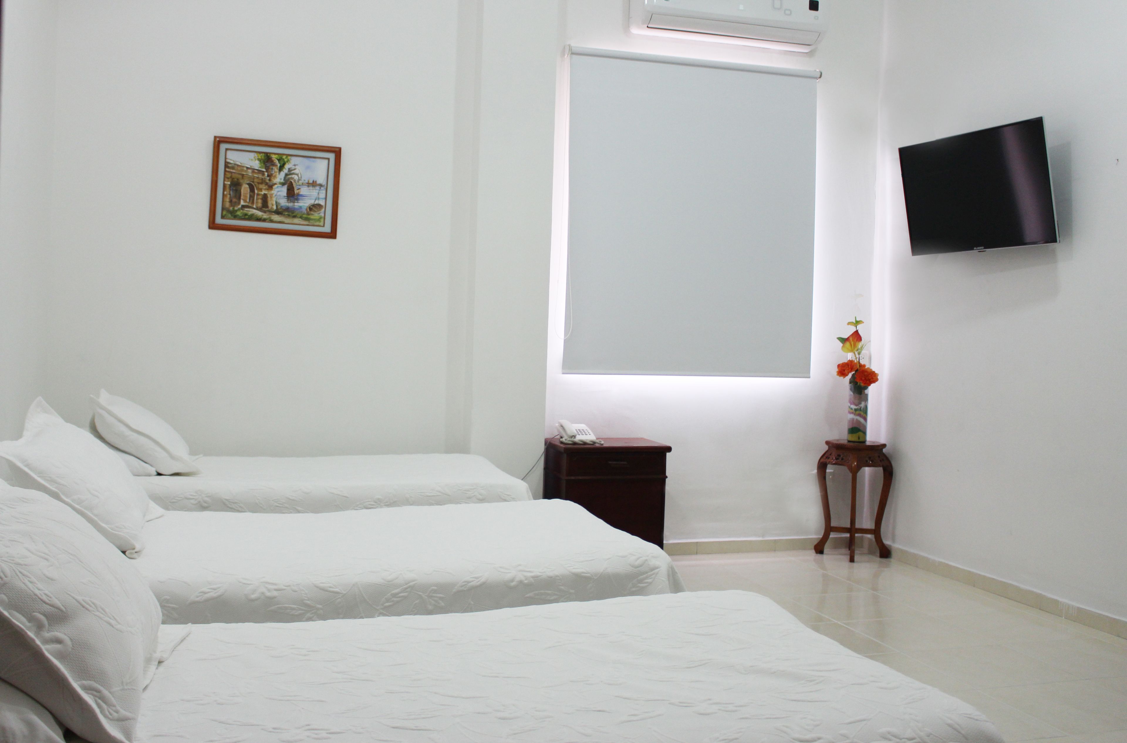 Habitación de hotel con dos camas, una mesita de noche pequeña con un teléfono, televisión montada en la pared, un cuadro enmarcado y una ventana con persianas.