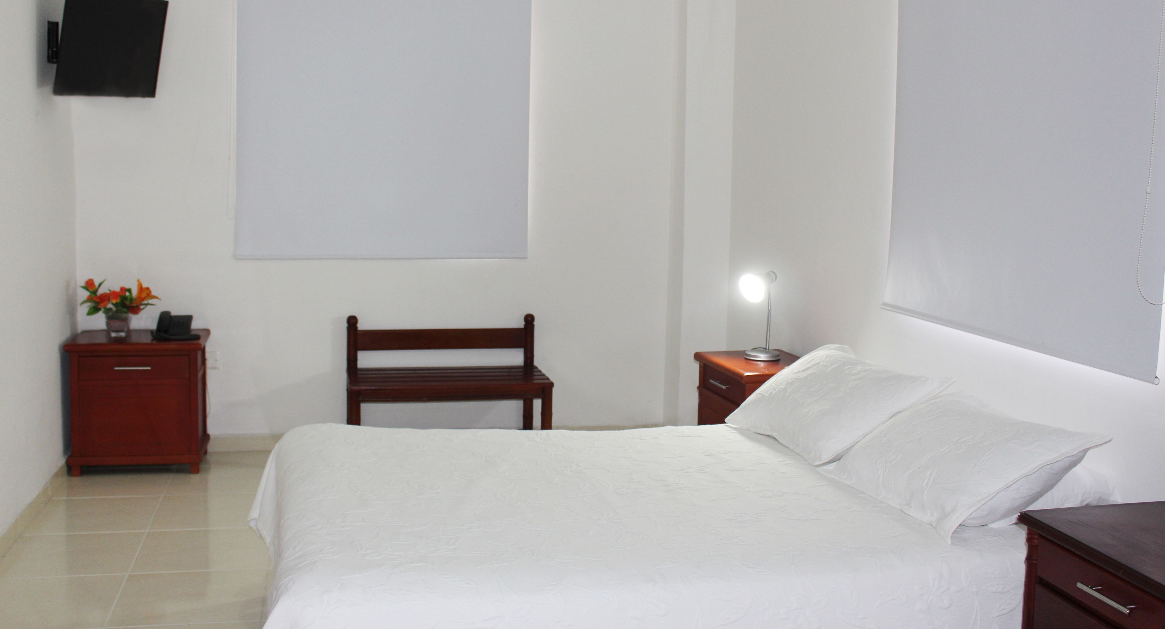 A neatly arranged bedroom with a bed, a wooden bench, a nightstand with a lamp, and a small table with a vase of flowers.