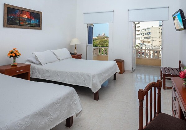 A hotel room with two beds, a desk, a chair, a nightstand, a lamp, a vase with flowers, a painting on the wall, and a balcony with a view of buildings and a dome structure. - Hotel Dorado Plaza Centro Histórico (Cartagena)
