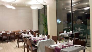A restaurant with tables set for dining, featuring white tablecloths, neatly arranged chairs, and hanging pendant lights.