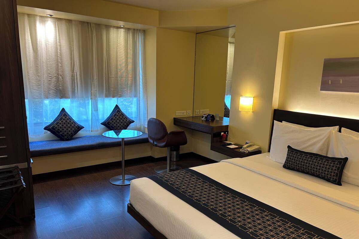 A hotel room with a bed, a desk, a chair, a window with curtains, a mirror, and a bedside table with a phone.