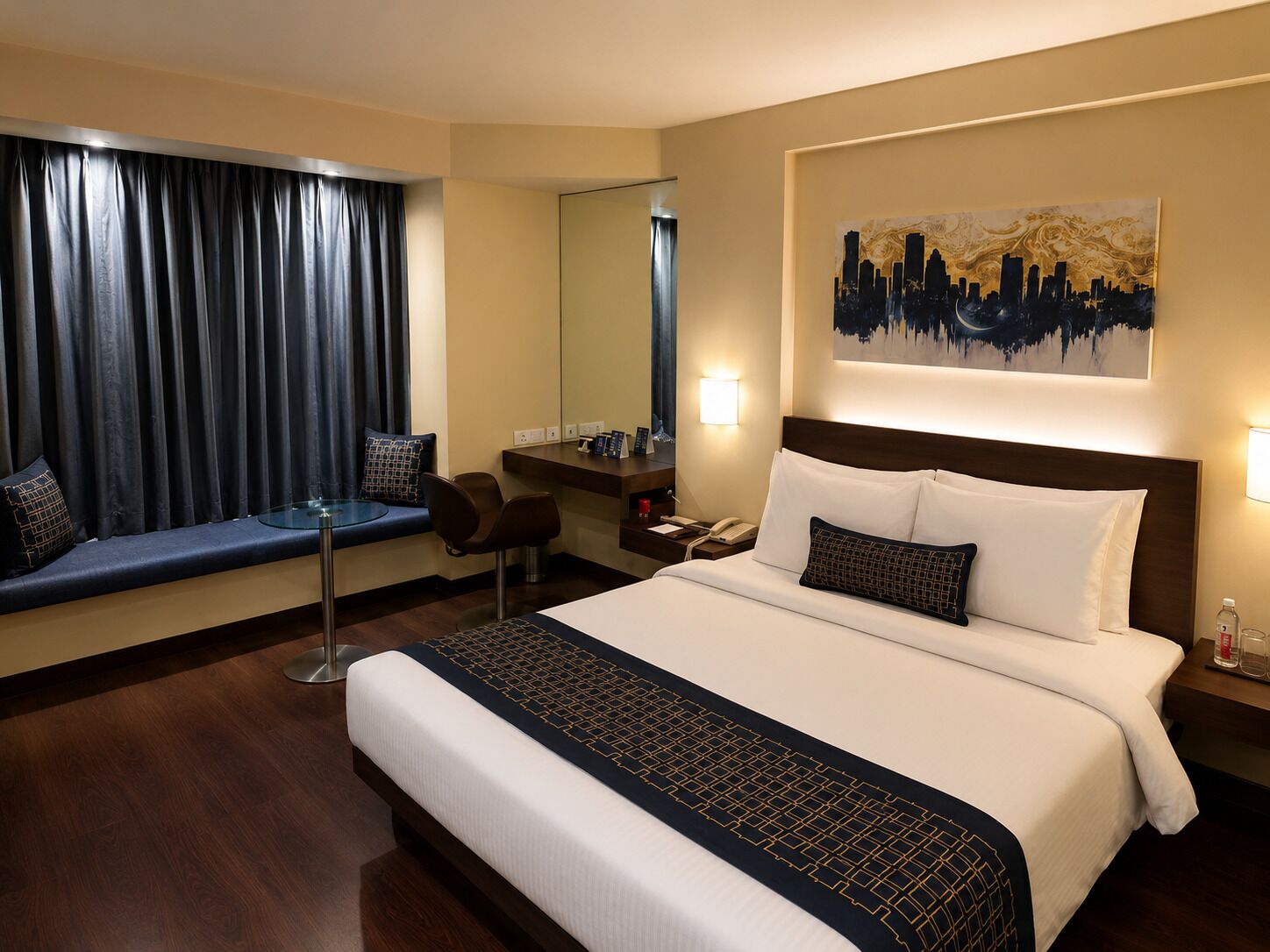 A hotel room with a large bed, a desk, a chair, a window with curtains, a wall-mounted painting, and a bedside table with a lamp.