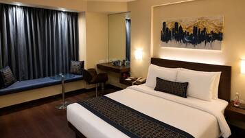 A hotel room with a large bed, a desk, a chair, a window with curtains, a wall-mounted painting, and a bedside table with a lamp.