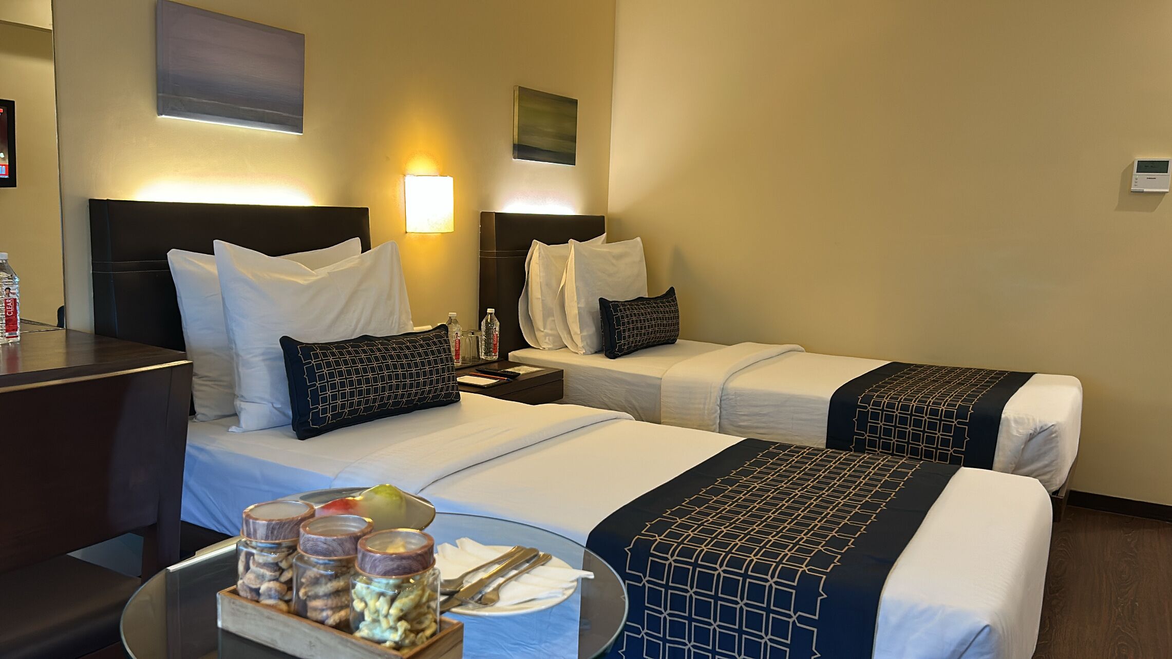 Hotelkamer met twee bedden, een klein tafeltje met een schotel noten, een fles water en een nachtkastje met een lamp.