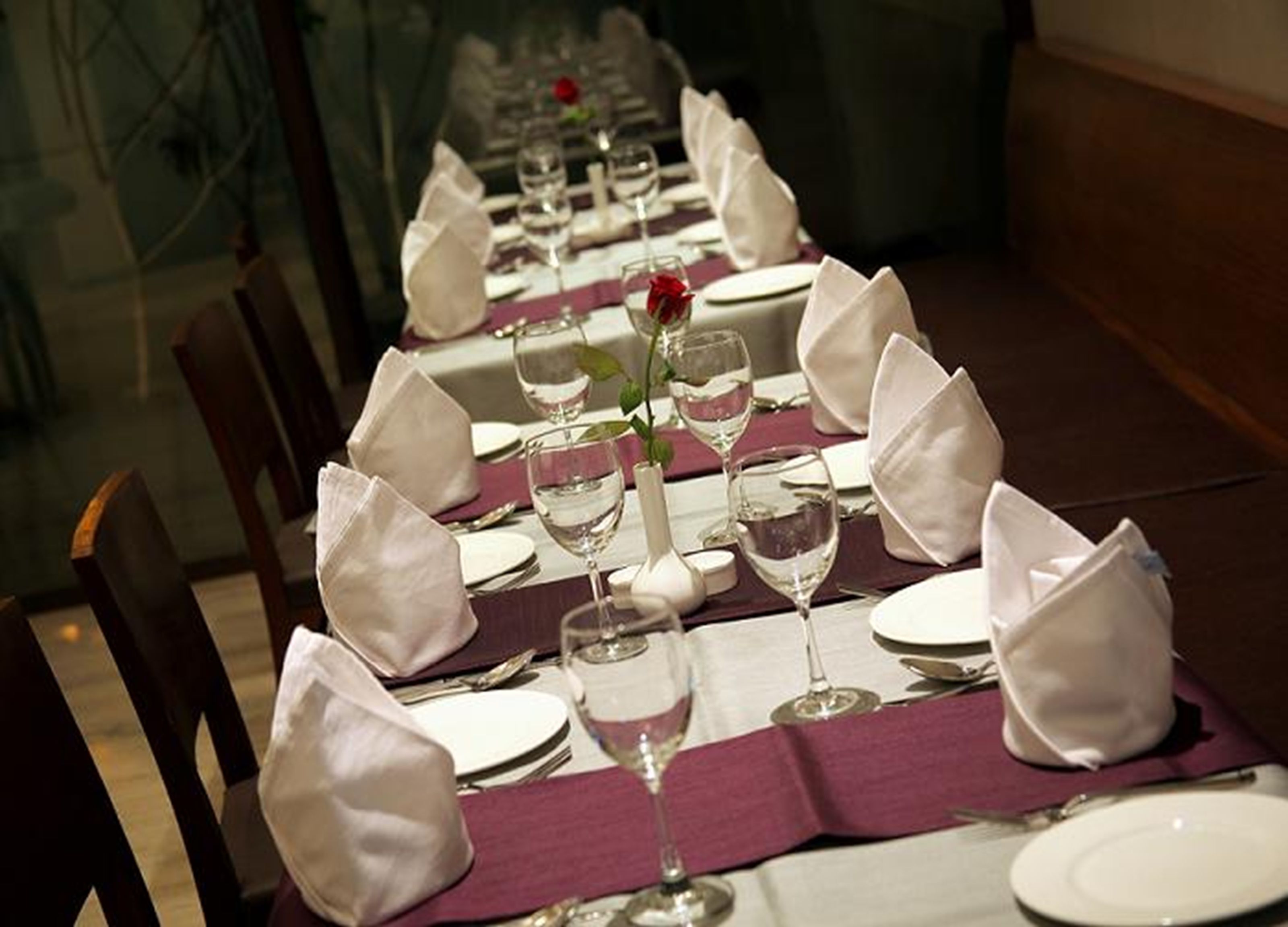 A long dining table set with white napkins, plates, and wine glasses, with a purple table runner and a single rose centerpiece.