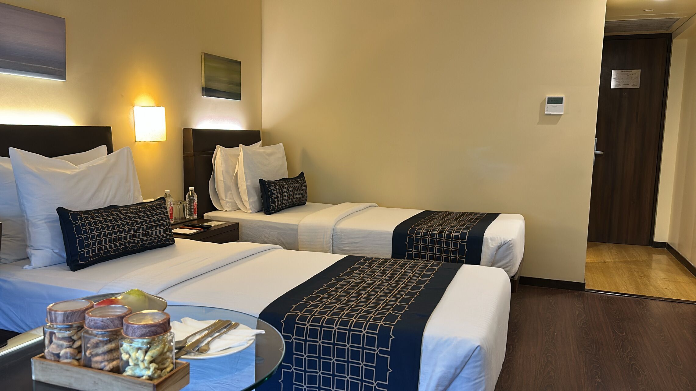 Ein Hotelzimmer mit zwei Betten, einem kleinen Tisch mit Glasplatte und einem Nachttisch mit Lampe.