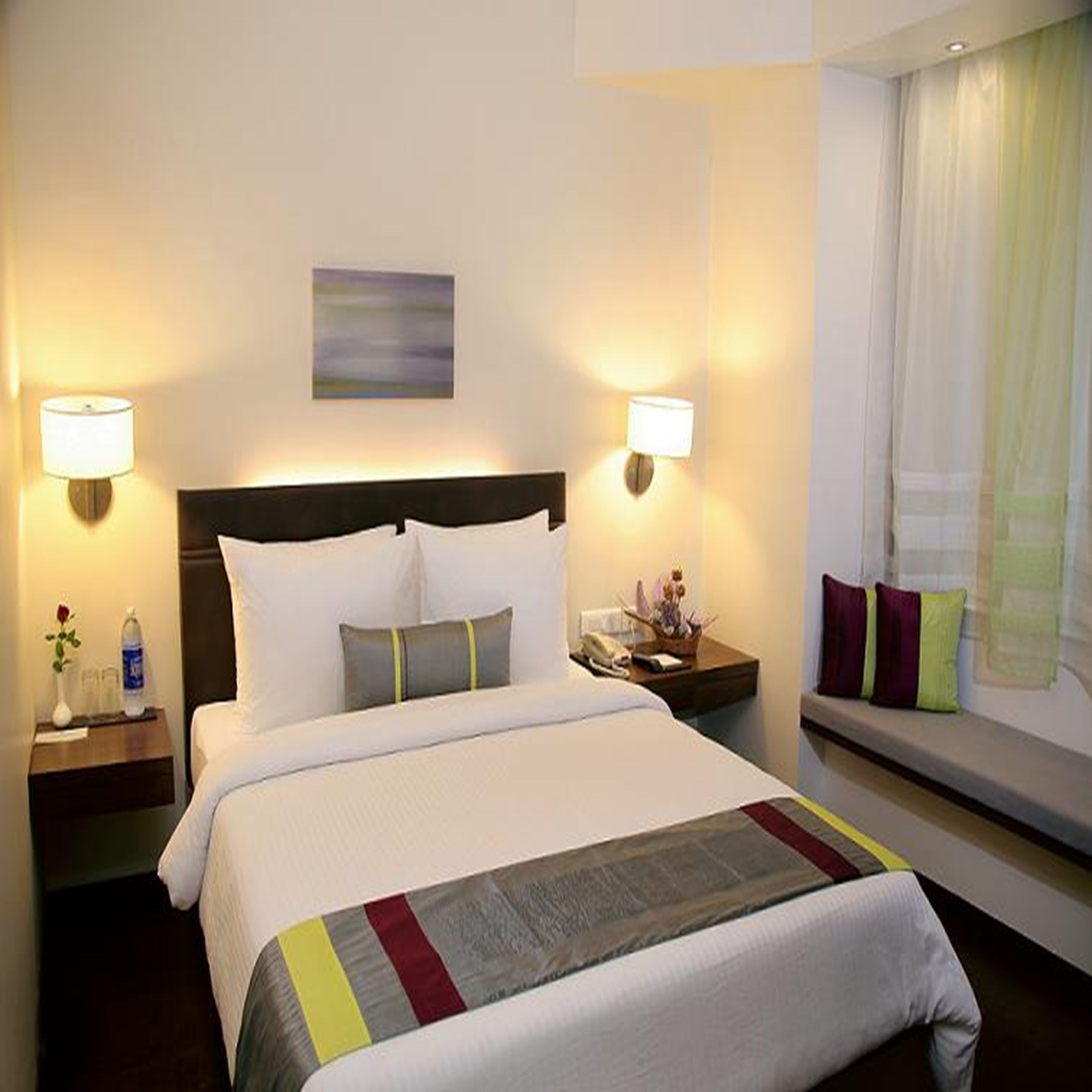 A hotel room with a large bed, two bedside tables, a nightstand, a window with curtains, and a wall-mounted lamp.