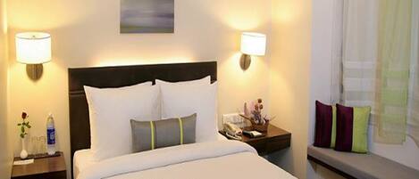A hotel room with a large bed, two bedside tables, a nightstand, a window with curtains, and a wall-mounted lamp.