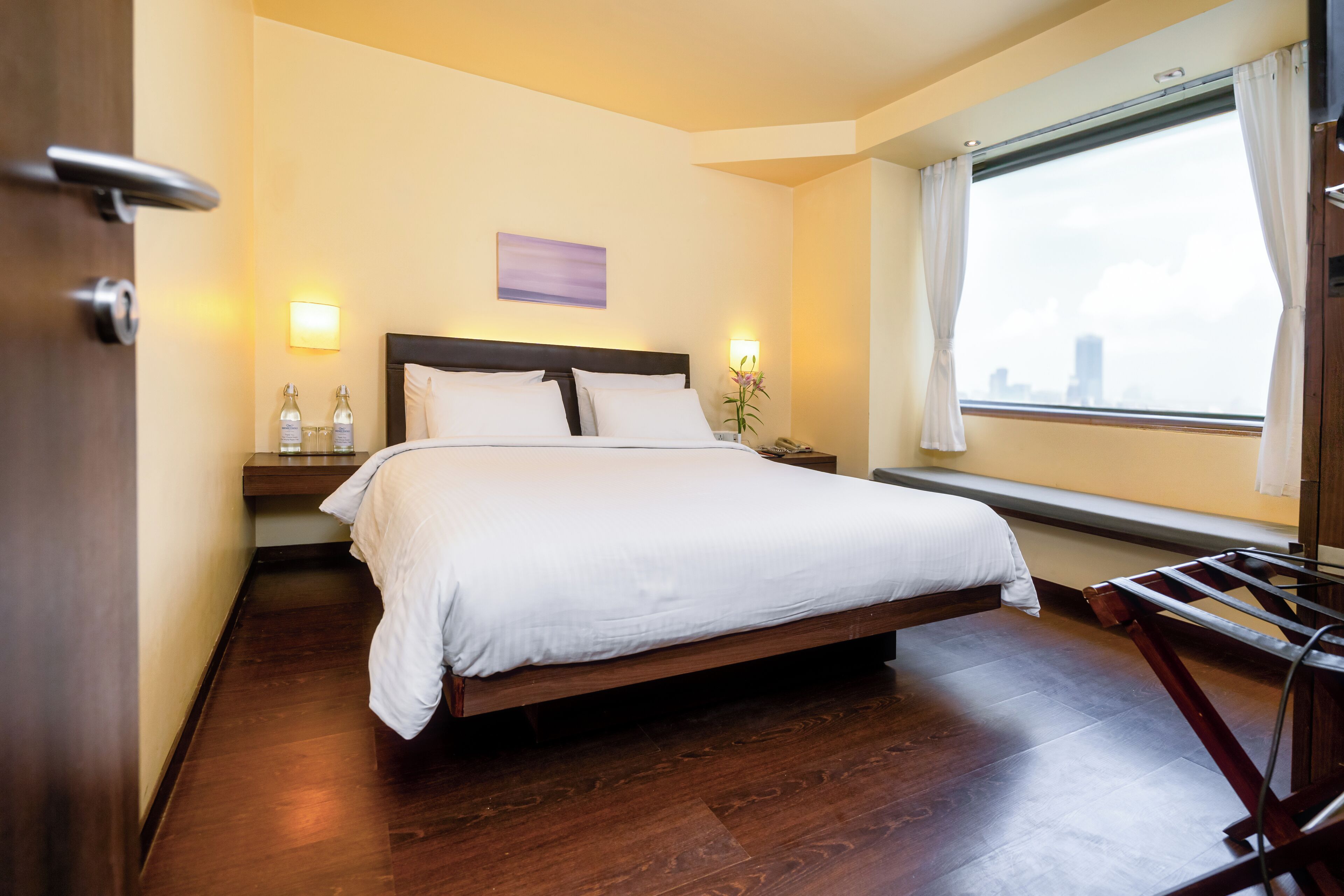 Camera d'albergo con un letto grande, pavimento in legno, un comodino con due bottiglie, una finestra con tende e vista sulla città.