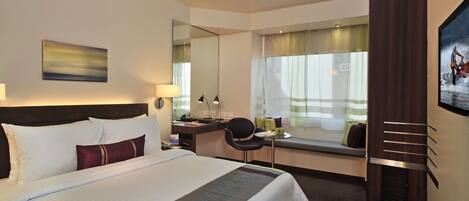 A hotel room with a large bed, a desk with a chair, a window with a view, and a painting on the wall.