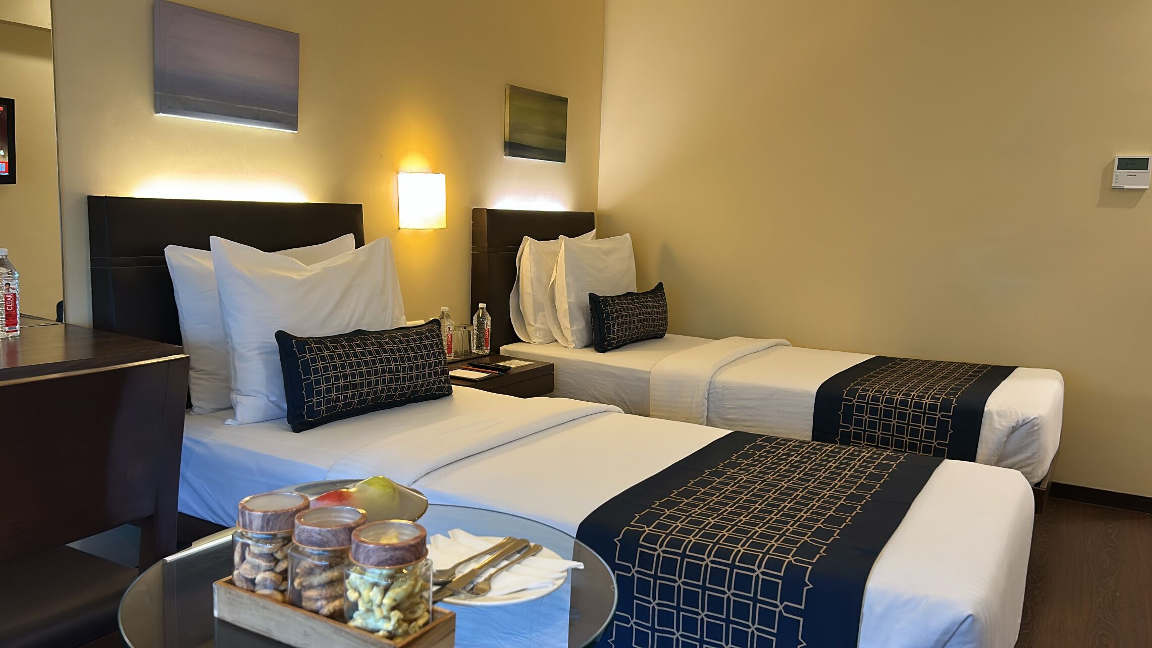 Habitación de hotel con dos camas, un escritorio, una mesita de noche, una mesa con frutero y frascos, y lámparas fijadas a la pared.