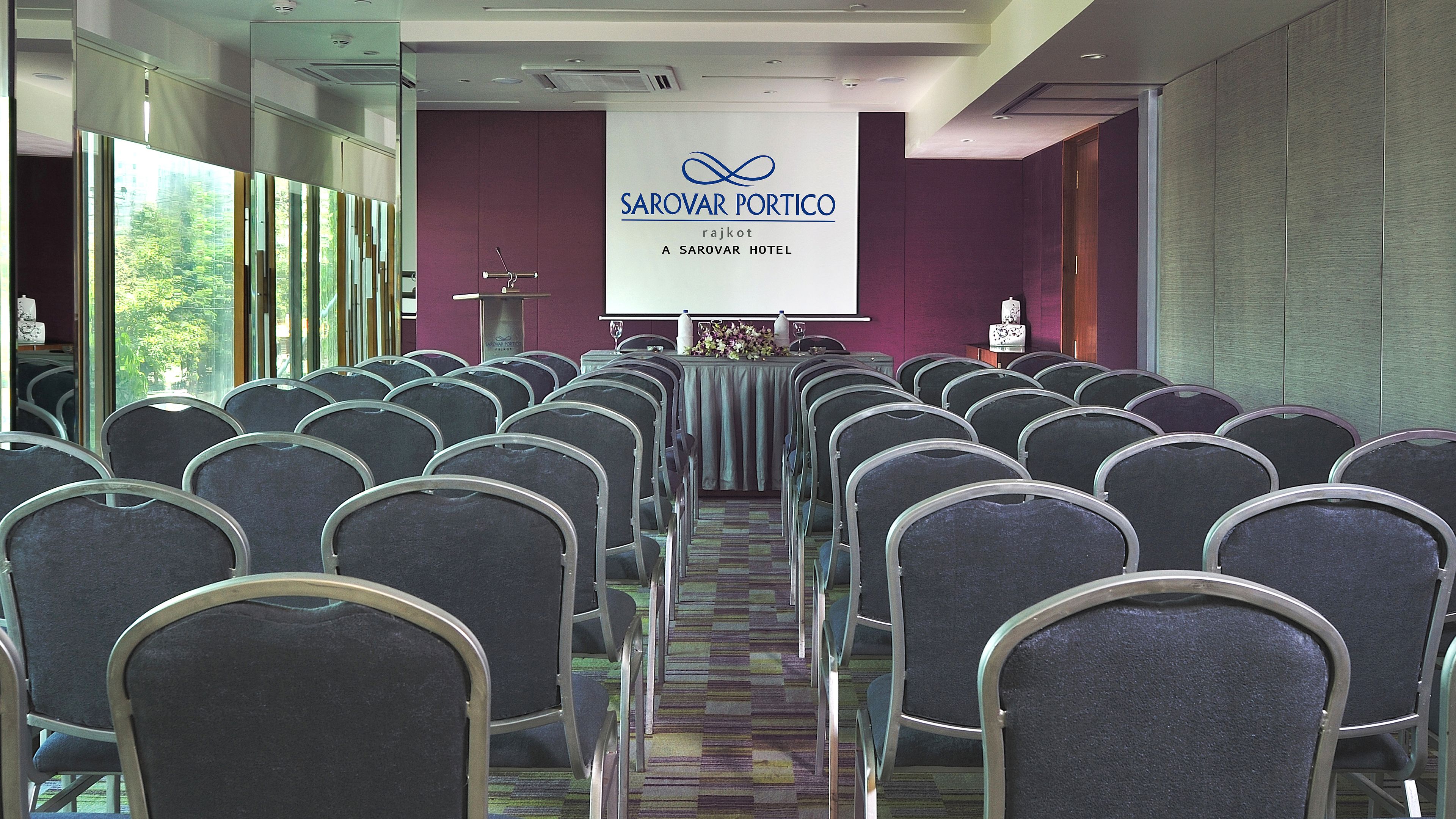 Un salón de conferencias con filas de sillas frente a un atril con un cartel que dice "SAROVAR PORTICO rajkot A SAROVAR HOTEL".