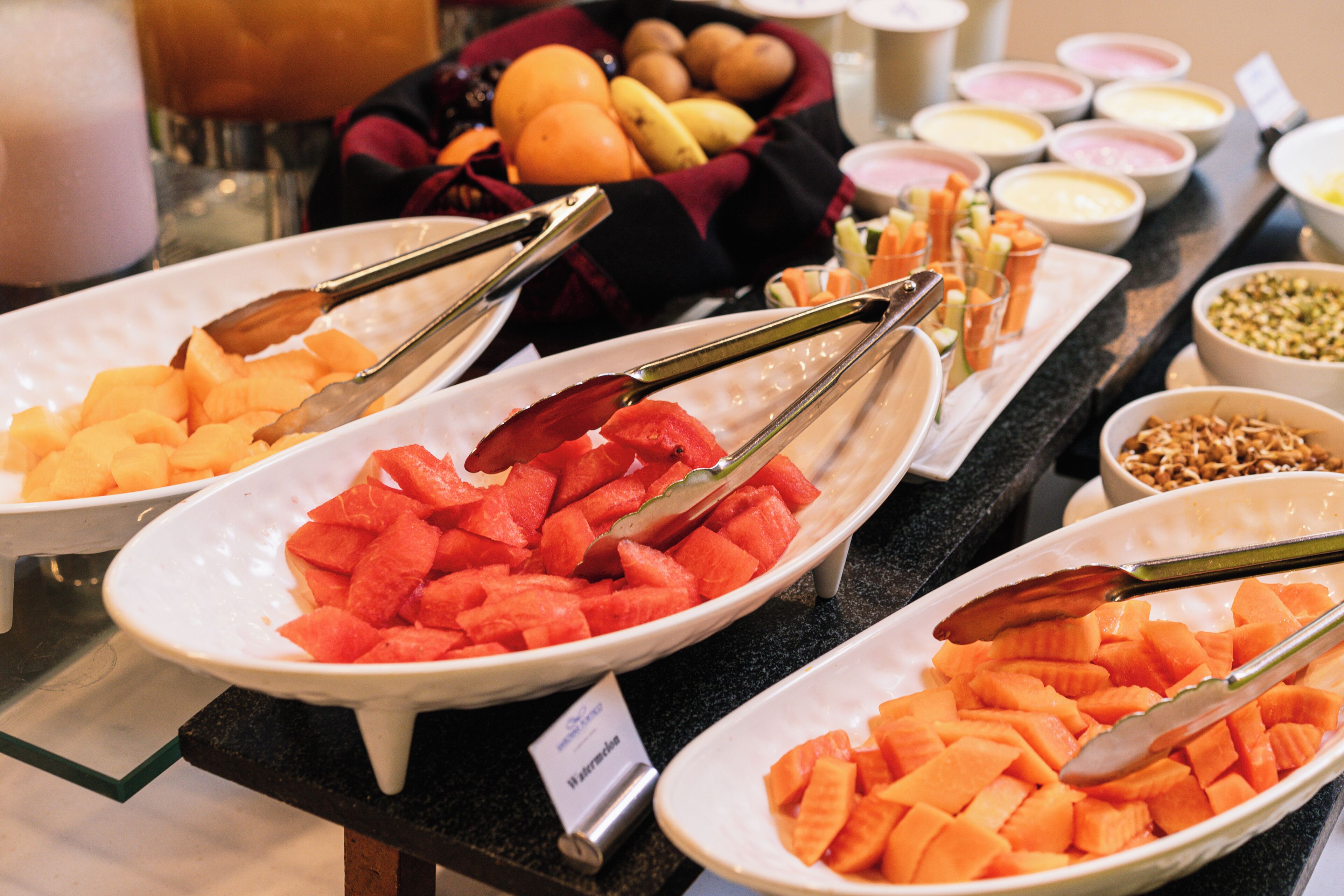 A buffet with various fruits and vegetables, including watermelon and carrots, arranged on white plates with tongs.
