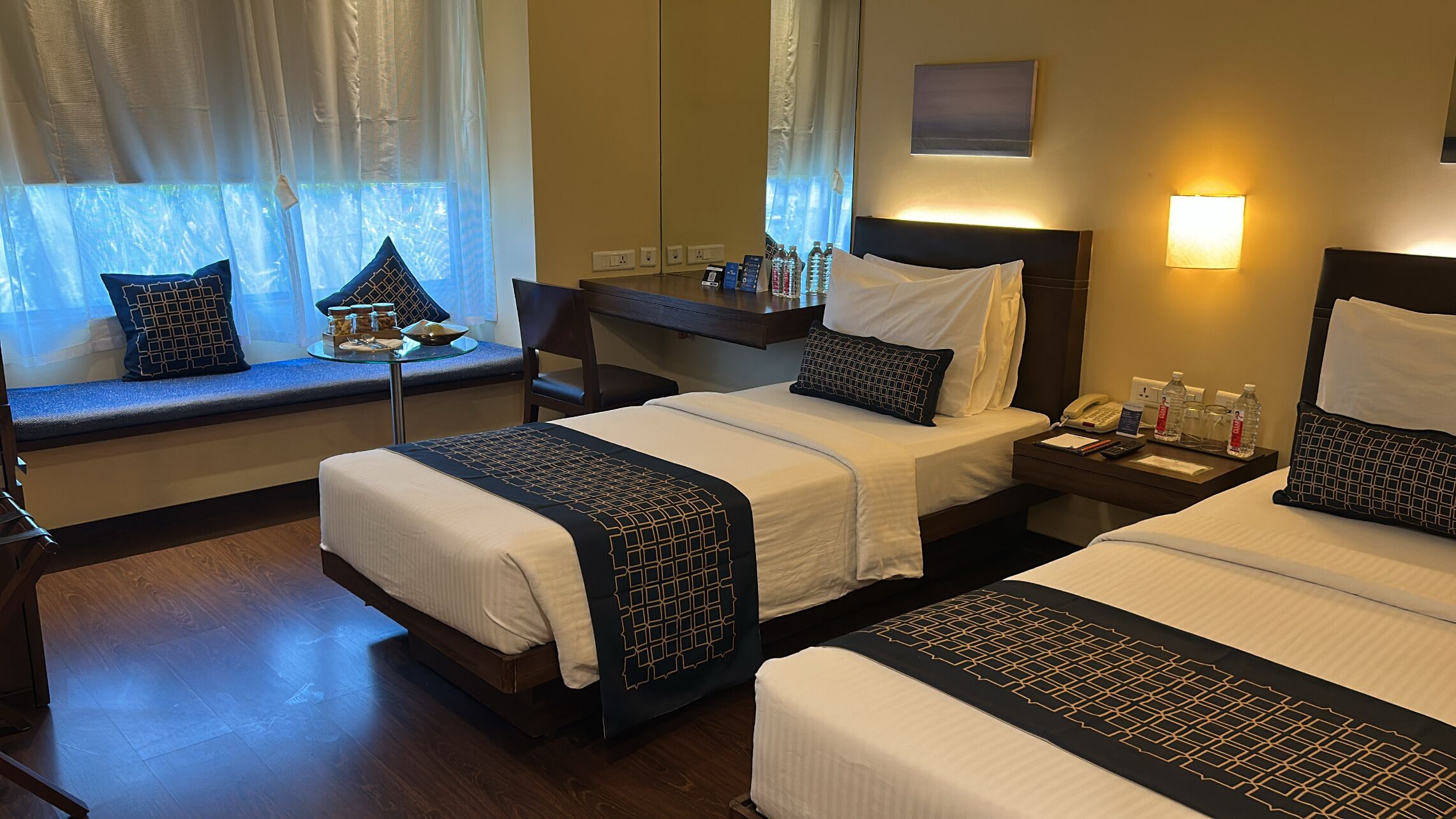A hotel room with two beds, a desk, a chair, a window with curtains, a nightstand with a phone and water bottles, and a wall-mounted lamp.
