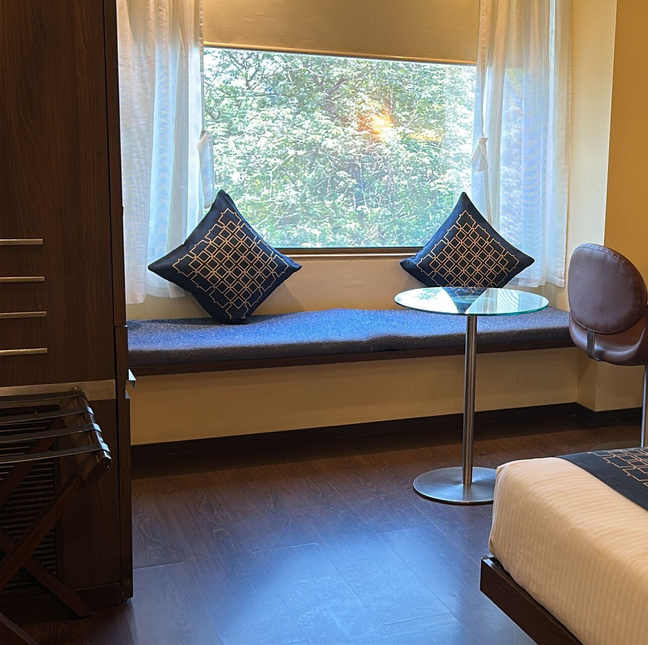A room with a window seat, two patterned pillows, a small round table, a chair, and a bed.