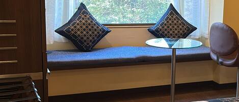 A room with a window seat, two patterned pillows, a small round table, a chair, and a bed.